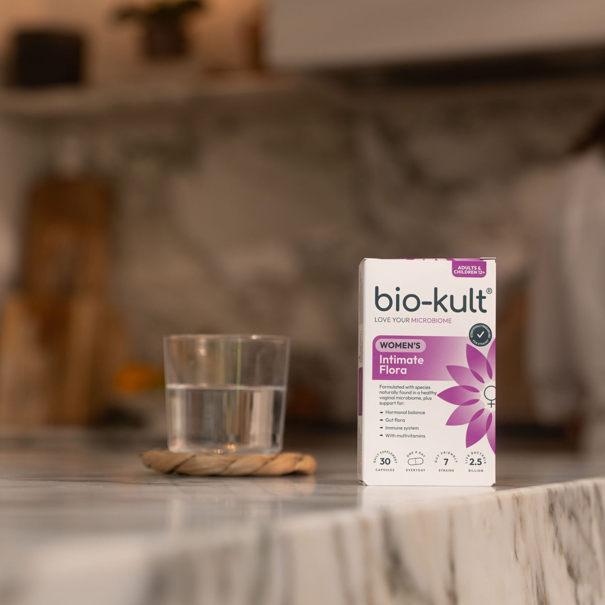 A box of bio-kult Women's Intimate Fibre dietary supplement stands on a marble countertop next to a glass of water in a blurred kitchen setting.