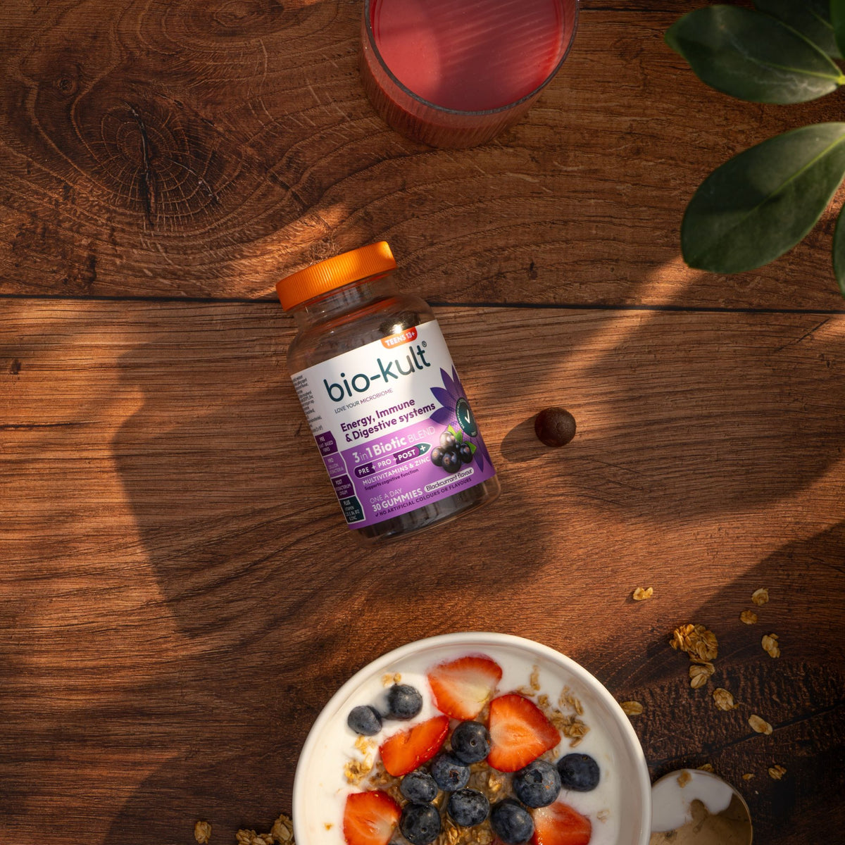 A bottle of bio-kult Energy Recharged Gummy Bears lies on a rustic wooden table next to a bowl of yogurt with strawberries and blueberries, with a glass of pink drink and green leaves in the background.