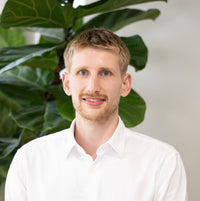 A man with short blond hair and light stubble, wearing a white button-down shirt, standing in front of a large green leafy plant against a light gray wall.